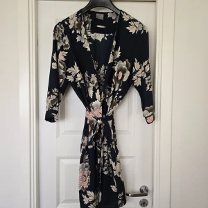 Blommig kimono från Vero Moda - Säljer en marinblå kimono från Vero Moda med vackert blommigt mönster i beige, rosa och grå toner. Kimonon har trekvartsärm och knytskärp i midjan. Perfekt att slänga över en enkel outfit för en snygg touch.