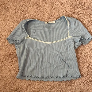 Ljusblå croppad topp från NA-KD - Säljer en ljusblå croppad topp från NA-KD med ribbad struktur och korta ärmar. Toppen har en söt spetsdetalj längs halsringningen och vågig kant nertill. Perfekt för vår och sommar! Tror den är från Misslisibels kollektion.