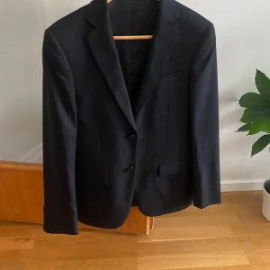 Svart kostym från Dressmann - Säljer en svart kostym från Dressmann i modellen Slim Fit, storlek 44 Short. Klassisk kavaj med två knappar och matchande kostymbyxor. Perfekt för bal eller fest. Snygg och stilren design.