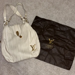 Beige vintage handväska från Louis Vuitton - Säljer en snygg vintage beige handväska från Louis Vuitton med guldiga detaljer och stort LV-logo framtill. Väskan har dubbla handtag, dragkedja på insidan och följer med en mörkbrun dustbag. Tillverkad i skinn med stilrena sömmar och exklusiv känsla.