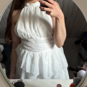 Vit halterneckklänning med volang - Säljer en vit klänning med halterneck och markerad midja. Klänningen har volangdetaljer vid kjolen och är ärmlös, perfekt för sommarens tillfällen. Snygg och luftig modell med romantisk känsla.