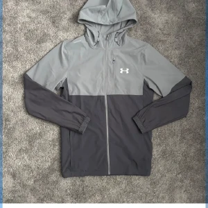 Grå och mörkgrå vindjacka från Under Armour - Snygg tvåfärgad vindjacka från Under Armour med huva och dragkedja. Jackan är grå upptill och mörkgrå nedtill, har långa ärmar och meshfoder på insidan. Perfekt för träning eller utomhusaktiviteter.
