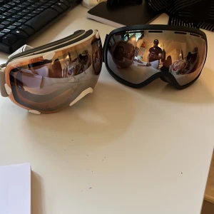 Skidglasögon med spegelglas och bred rem - Säljer två par skidglasögon med spegelglas. Ena paret har svart ram och svartvit rem med Crosportwear-tryck, det andra har beige ram och beigevit rem med korsmönster. Båda har breda elastiska band och ventilerad ram. Ops!! Den vita har flera repor