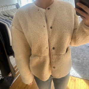 Beige teddyjacka från H&M - Mysig beige teddyjacka från H&M med rund halsringning och tryckknappar framtill. Jackan har två fickor och är helfodrad. Perfekt för lager-på-lager och ger en trendig look till vardagsoutfiten.
