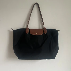 Svart axelväska med bruna detaljer - Stilren svart axelväska från Longchamp med bruna skinnhandtag och lock med guldfärgad knapp. Väskan är rymlig och har klassisk design, perfekt för att bära med sig allt man behöver.