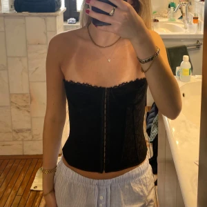 Svart tubtopp korsett - Säljer en svart tubtopp i korsettmodell med dragkedja framtill. Toppen är axelbandslös och har en lätt glansig yta med diskreta sömmar som ger en snygg form. Perfekt att styla till både jeans och kjol.