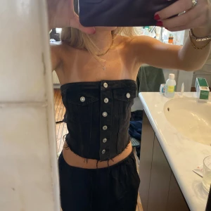 Svart jeanskorsett med knappar - Säljer en svart korsettopp i jeanstyg med silverfärgade knappar framtill och dekorativa fickor. Toppen är axelbandslös och har en tight passform som framhäver midjan. Perfekt för att skapa en edgy look.