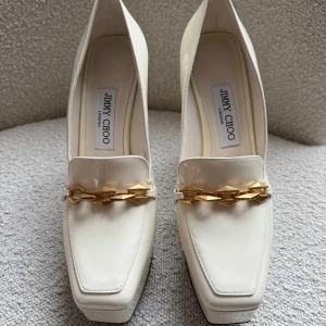Vita platåpumps från Jimmy Choo - Säljer ett par eleganta vita platåpumps från Jimmy Choo med fyrkantig tå och chunky klack. Cremefärg. Skorna har en snygg guldfärgad kedjedetalj framtill och är tillverkade i blankt syntetmaterial. Perfekta för dig som vill sticka ut med elegans. 100% äkta. Inköpta i Jimmy Choo butiken i Paris. Använt dem enbart 2-3 ggr. 
