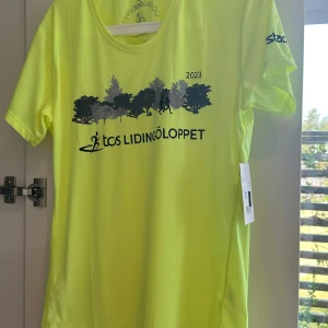 Gul Lidingöloppet t-shirt från SOC - Fräsch gul t-shirt från SOC med Lidingöloppet 2023-tryck framtill och logga på ryggen. T-shirten har korta ärmar och är gjord i ett lätt, syntetiskt material som passar perfekt till träning. Snygg och sportig look med tryck i svart och blått.