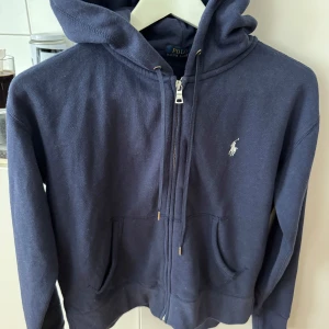 Mörkblå hoodie från Polo Ralph Lauren - Mörkblå hoodie från Polo Ralph Lauren med dragkedja och huva. Klassisk broderad logga på bröstet och två fickor framtill. Perfekt för en avslappnad stil.