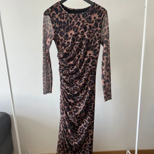 Leopardmönstrad långklänning med rynkade detaljer - Säljer en långärmad långklänning med leopardmönster i svart och beige. Klänningen har rynkade detaljer längs sidan som ger en snygg siluett. Tillverkad i ett tunt, stretchigt syntetmaterial. Perfekt för dig som vill sticka ut!
