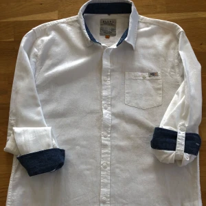 Vit skjorta med blå detaljer från Blend - Vit långärmad skjorta från Blend med kontrastblå insida på krage och ärmslut. Klassisk knappslå och bröstficka. Perfekt för en stilren look med en twist.