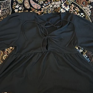 Svart klänning med knyt framtill - Svart klänning med två knytningar framtill och lätt puffade ärmar. Tyget är strukturerat och klänningen har en lös passform under bysten. Perfekt för dig som gillar en enkel men ändå detaljrik look.