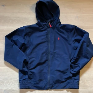 Mörkblå vindjacka från Polo Ralph Lauren - Snygg mörkblå vindjacka från Polo Ralph Lauren med huva och dragkedja. Jackan har en röd logotyp på bröstet och elastiska muddar. Perfekt för blåsiga dagar.