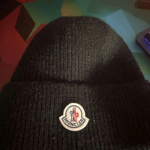 Svart ribbad mössa från Moncler - Snygg svart ribbad mössa från Moncler med deras ikoniska logotyp framtill. Perfekt för att hålla värmen under kyliga dagar. Stilren design som passar till det mesta.