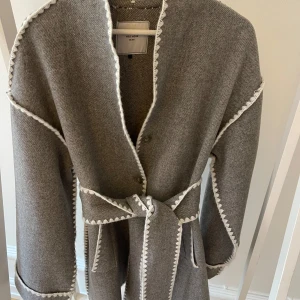 Grå kappa från Neo Noir - Snygg grå kappa från Neo Noir med vita broderade detaljer längs kanterna. Kappan har ett bälte i midjan och långa ärmar. Perfekt för en stilren look.