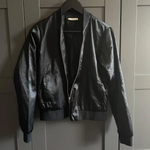 Svart bomberjacka - Svart bomberjacka i glansigt material från Pieces med dragkedja framtill. Jackan har ribbade muddar vid ärmslut och nederkant, samt fickor framtill. 
