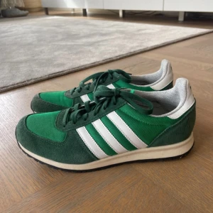 Gröna sneakers från Adidas - Snygga gröna sneakers från Adidas med vita ränder och snörning. Perfekta för en avslappnad stil. Skorna har en bekväm passform och en klassisk design som aldrig går ur tiden.