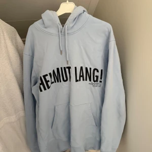 Ljusblå hoodie från Helmut Lang - Säljer en stilren ljusblå hoodie från Helmut Lang med svart texttryck på framsidan. Tröjan har en klassisk känguruficka och justerbar huva med dragsko. Perfekt för en avslappnad stil. Hör av dig vid intresse!