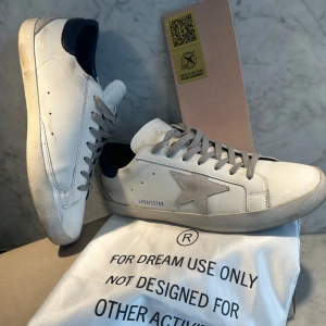 Golden goose  - Snygga vita sneakers från Golden Goose med en ikonisk stjärndetalj på sidan. Skorna har en  mörkblå häl och grå snörning. Jätte bra skick har bara en liten skada på baksidan av sulan. Bara att  höra av er vid frågor. Box, påse och allt annat ingår. Är också öppen för byten.