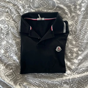 Svart pikétröja från Moncler - Snygg svart pikétröja från Moncler med klassisk krage och vita detaljer på kragen och ärmsluten. Perfekt för en stilren look. Tröjan har en broderad logotyp på bröstet.