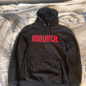 Svart hoodie från Hoodrich - Säljer en svart hoodie från Hoodrich med röd logga på bröstet. Den har en klassisk känguruficka och justerbar huva med snören. Perfekt för en avslappnad stil.