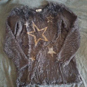 Grå fluffig tröja med stjärnor från H&M - Säljer en supermjuk och fluffig grå tröja från H&M med stora stjärnor i olika storlekar på framsidan. Perfekt för att mysa i under kyliga dagar. Tröjan har långa ärmar och en bekväm passform. För barn