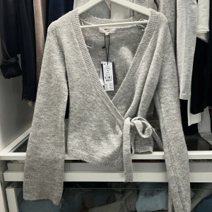 Grå omlottkofta från Gina Tricot - Säljer en ljusgrå omlottkofta från Gina Tricot i ullblandning. Koftan har en knytning i midjan och långa ärmar. Den är aldrig använd lappen kvar! Storlek S. Skriv om du vill se bilder hur den sitter på💗💞💞