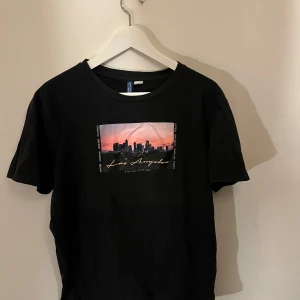 Svart t-shirt med Los Angeles-tryck - Snygg svart t-shirt med ett coolt tryck av Los Angeles skyline i solnedgång. Perfekt för en avslappnad stil. Kortärmad och i mjukt material.