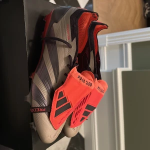 Adidas Predator fotbollsskor - Snygga Adidas Predator fotbollsskor i grått och rött med svarta detaljer. Butikpris är 3100kr Skorna har snörning och en strömlinjeformad design för optimal prestanda på planen. Perfekta för fotbollsentusiaster som vill ha stil och funktionalitet.  Använda en gång i 30 min bara, säljer pga fel storlek och kan inte lämna tillbaka! 