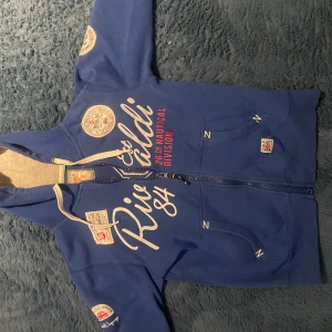 Blå huvtröja från Ralph Lauren - Säljer en blå huvtröja från Rivaldi med dragkedja och tryck i vitt och rött. Tröjan har en stor 26 på ryggen och flera märken på ärmarna. Perfekt för en avslappnad stil.