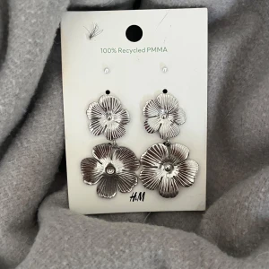 Silvriga blommiga örhängen från H&M - Snygga silvriga örhängen från H&M i form av blommor. Tillverkade av 100% återvunnen PMMA, dessa örhängen är både stiliga och miljövänliga. Perfekta för att ge en glittrig touch till din outfit. Helt nya! Och helt oanvända!