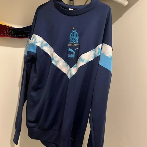 Marseille Tracksuit från Puma - Marseille Tracksuit/Säljer en blå långärmad tröja från Puma med vit och ljusblå detalj över bröstet. Tröjan har en rund hals och klubbmärke på bröstet. Perfekt för sportiga tillfällen.