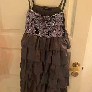 Svart klänning med paljetter - Snygg svart klänning med glittriga paljetter upptill och volanger nedtill. Axelbandslös design med elastisk överdel för en bekväm passform. Perfekt för en festlig kväll!