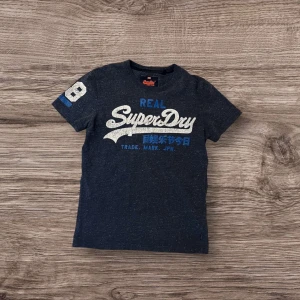Mörkblå t-shirt från SuperDry - Säljer en mörkblå t-shirt från SuperDry med tryck i vitt och blått på framsidan. T-shirten har korta ärmar och ett nummertryck på ena ärmen. Perfekt för sommaren🙌
