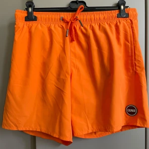 Orange badshorts från italienska Colmar - Snygga orange badshorts från Colmar med elastisk midja och snörning. Perfekta för sommarens strandhäng. Shortsen har en diskret logga på benet och är tillverkade i ett lätt och snabbtorkande material.