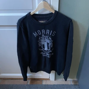 Svart tröja från Morris - Säljer en svart tröja från Morris med ett tryck på framsidan och en liten flagga på ärmen. Tröjan har långa ärmar och en rund halsringning. Perfekt för en stilren look. Storlek M