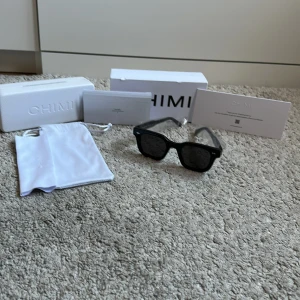 Svarta solglasögon från Chimi - Snygga svarta solglasögon från Chimi med en klassisk fyrkantig form. De har mörka linser och kommer med originalförpackning och en vit förvaringspåse. Perfekta för att ge en stilren look. De är använda, har några skråmor men är i riktigt bra skick. Priset är inte hugget i sten!