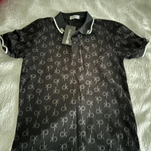 Svart kortärmad pikétröja från Calvin Klein - Snygg svart pikétröja från Calvin Klein med ett diskret mönster av CK-loggan över hela tyget. Tröjan har vita detaljer på kragen och ärmsluten, vilket ger en stilren kontrast. Perfekt för en avslappnad men ändå stilfull look. Passar m