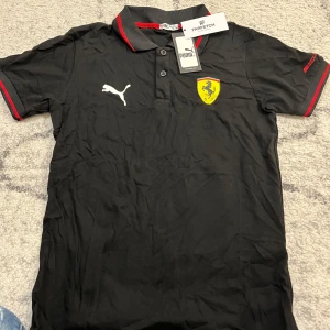 Svart pikétröja från Puma med Ferrari-logga - Snygg svart pikétröja från Puma med röd detaljering och Ferrari-logga på bröstet. Tröjan har en klassisk krage och knappar framtill. Perfekt för bilentusiaster och stilmedvetna. M