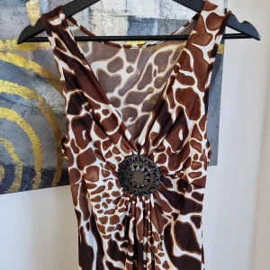 Brunmönstrad klänning med dekorativ detalj - Snygg klänning med ett djurinspirerat mönster i brunt och vitt. Klänningen har en iögonfallande dekorativ detalj framtill och är ärmlös. Perfekt för en unik stil.