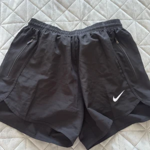 Svarta träningsshorts från Nike - Säljer ett par svarta träningsshorts från Nike med elastisk midja och dragkedjefickor. Perfekta för löpning eller gymmet. De har en klassisk Nike-logga i vitt på benet.