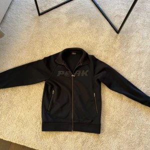 Svart jacka från Peak Performance - Säljer en svart jacka från Peak Performance med dragkedja och broderad logga på bröstet. Jackan har långa ärmar och två fickor med dragkedja. Perfekt för en sportig look!