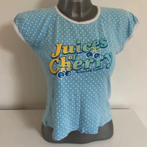 Blå prickig t-shirt med tryck - Säljer en blå t-shirt med vita prickar och ett färgglatt tryck 'Juices of Cherry' på framsidan. T-shirten har korta ärmar och en rund halsringning med vit kant. 