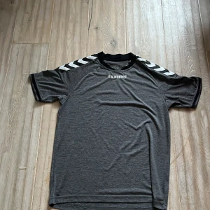 Grå kortärmad tröja från Hummel - Säljer en grå kortärmad tröja från Hummel med vita detaljer på axlarna. Perfekt för träning eller vardagsbruk. Tröjan har en bekväm passform och är tillverkad i ett lätt material. Säljer för att den är för stor  passar även xs