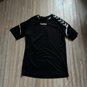 Svart sporttröja från Hummel - Säljer en svart sporttröja från Hummel med vita detaljer och korta ärmar. Perfekt för träning eller fotboll. Tröjan har Hummels ikoniska pilar på ärmarna och en bekväm passform.
