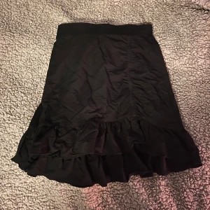 Svart kjol med volang - Snygg svart kjol med volangdetalj i nederkant. Perfekt för en stilren look. Kjolen har en normal passform och är gjord i ett mjukt material som ger en bekväm känsla.