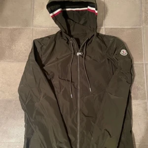 Moncler windbreaker - Snygg grön vindjacka från Moncler med dragkedja och huva. Jackan har Moncler-logga på ärmen och snörning vid huvan. Perfekt för blåsiga dagar. Kan mötas i stockholms området, annars frakt! Jackan är äkta, har tyvärr inget kvitto.