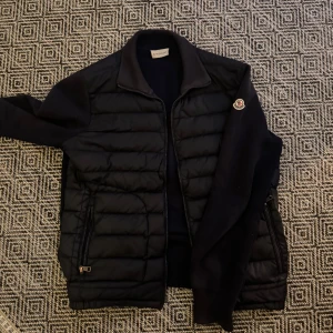 Mörkblå cardigan från Moncler - Snygg svart cardigan från Moncler med dragkedja och quiltad design. Perfekt för att hålla värmen med stil. Cardigan har en klassisk look och passar bra till olika outfits. Lite solblekt och moncler tagen är förstörd pm för mer info och bilder litet hål vid högre ärmen 