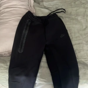 Nike tech byxor - Säljer ett par svarta mjukisbyxor från Nike med dragkedja på sidorna och snörning i midjan. Perfekta för en avslappnad stil eller träning. Byxorna har en stilren design med diskret logga.